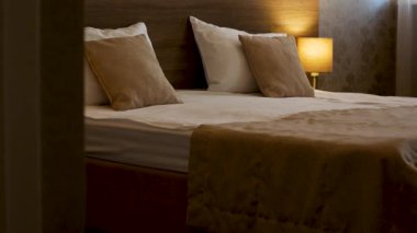 Geniş yatağı ve yumuşak yastıkları olan rahat bir yatak odasının içi. Yatağın yanında sıcak ışık ve penceresinde beyaz perdeler olan bir lamba var. Yatak odasındaki sıcak akşam atmosferi, konfor ve...