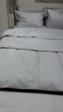 Gri yataklı bir yatak, battaniye ve yastıklar, lambalı bir dolabın yanında. Yatak odasının içi..