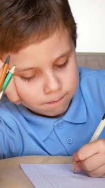 6-7 yaşlarında sevimli bir çocuk defterine kalem koyup başını dinlendiriyor. Yanındaki camda renkli kalemler var. Karantina sırasında uzaklık öğrenme