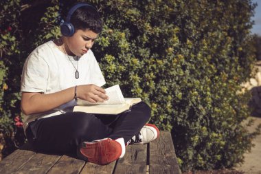 young man turning the pages of the book he is reading, sitting on the wooden bench in the garden of the country house, on the outskirts of the city,