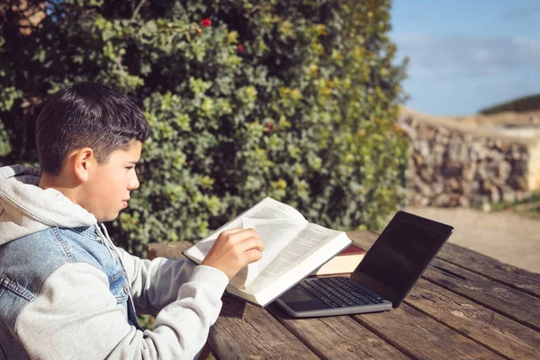 young latinstudent turns the page of a book in front of a laptop, outdoors in an urban park, doing high school study work.