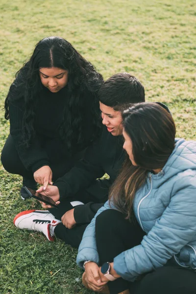 three people of hispanic-latino ethnicity sitting on the green grass of a park, with smart device. relaxed and comfortable as they entertain themselves with their screens, browsing social networks