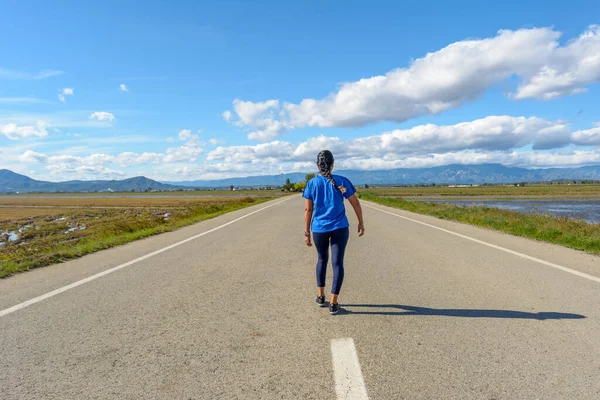 A person walks down a straight road with mountains and a clear sky ahead, latina woman walking along lonely road center in the Ebro Delta natural park, Tarragona, Catalonia, Spain,