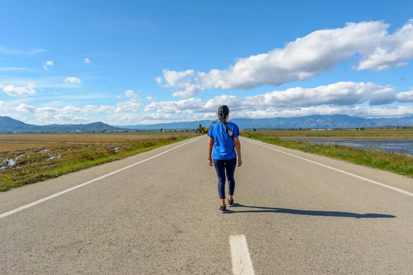 A person is walking down an open road surrounded by nature under a cloud-streaked blue sky, back view of latina woman dressed in blue walking down the road in the Ebro Delta natural park, Tarragona