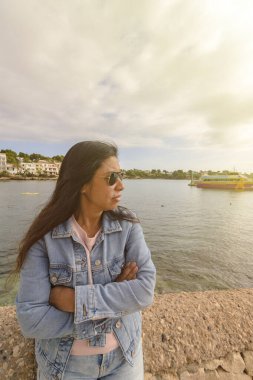 Kot ceketli ve güneş gözlüklü Latin kadın bulutlu bir gökyüzünün altındaki suya, porto petrolüne, mallorca balear adalarına, İspanya 'ya bakar..,