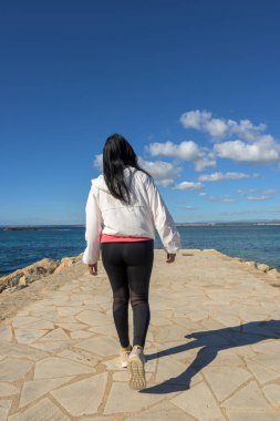 Okyanus kıyısındaki taştan bir yolda tek başına yürüyen bir kadın, bulutlarla birlikte açık mavi bir gökyüzünün altında, Colonia San Jordi, Balear Adaları, Mayorka, İspanya 'da deniz kenarında güzel bir Latin kadın.