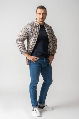 Ekose gömlekli, mavi tişörtlü ve kalçasında kot pantolon olan atletik bir adam. Beyaz arkaplanda poz veren model.