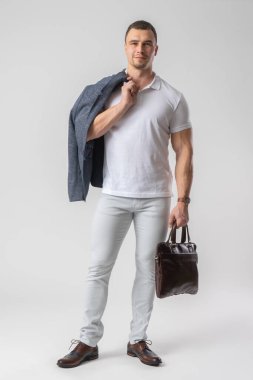 Günlük hafif giysiler içinde atletik yapılı bir adam elinde deri bir çanta ve bir ceket tutuyor. Beyaz arkaplanda poz veren model.