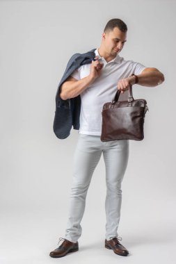 Sıradan giyinmiş bir adam saatine bakar ve elinde deri bir evrak çantası ve ceket tutar. Beyaz arkaplanda poz verme