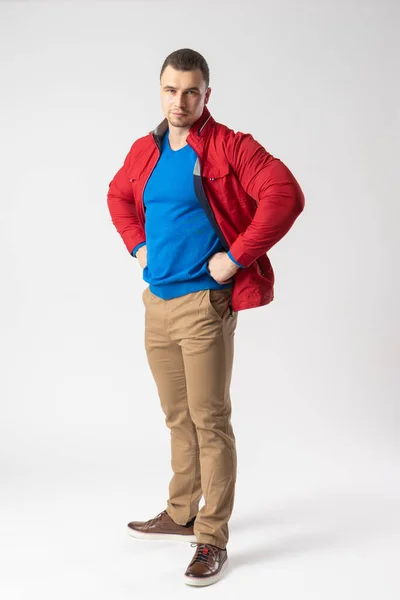 赤のジャケット 青のTシャツ ベージュのパンツを着た運動用の建物の男がAkimbo立っています 白地にポーズをとるモデル — ストック写真