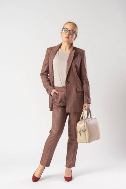 Gözlüklü genç sarışın bir kadın ve elinde bir kadın çantasıyla sıradan bir ofis elbisesi. Stüdyoda beyaz arka planda poz veren bir model..