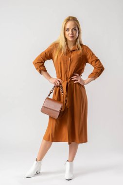 Zarif, kahverengi elbiseli, küçük kahverengi deri çantalı sarışın bir kadın. Stüdyoda beyaz arka planda poz veren bir model..