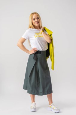 Şık, genç, rahat bir etek ve beyaz bir tişört giymiş genç sarışın bir kadın, omzunun üzerinden atıyor, açık yeşil deri bir ceket. Stüdyoda beyaz arka planda poz veren bir model..