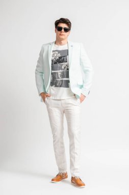 Açık renk ceketli ve güneş gözlüklü genç siyah saçlı bir adam beyaz arka planda stüdyoda uzun süre poz veriyor.