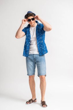 Kısa kollu, kısa kollu, beyaz tişörtlü, kot şortlu, şapkalı, güneş gözlüklü genç adam beyaz arka planda poz verirken ya güneş gözlüğü takıyor ya da çıkarıyor.
