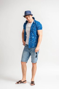 Kısa kollu, beyaz tişörtlü, kot şortlu, siyah saçlı genç bir adam elinde güneş gözlüğü tutuyor ve beyaz bir arka planda stüdyoda uzun süre poz veriyor.