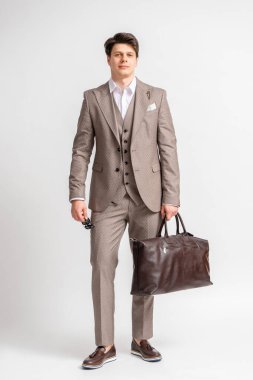 Siyah saçlı, iş elbiseli, güneş gözlüklü, seyahat ve iş seyahatleri için büyük deri çantalı, beyaz bir arka planda stüdyoda poz veren genç bir adam.
