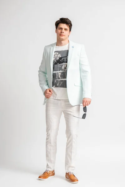 白い背景の上にスタジオで長さをポーズカジュアルライトカラーのジャケットとサングラスの若いダークヘアの男 — ストック写真