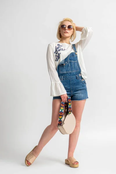 stock image Young blonde woman in casual summer denim shorts and a white rustic blouse with a handbag in her hand and sunglasses posing on a white background
