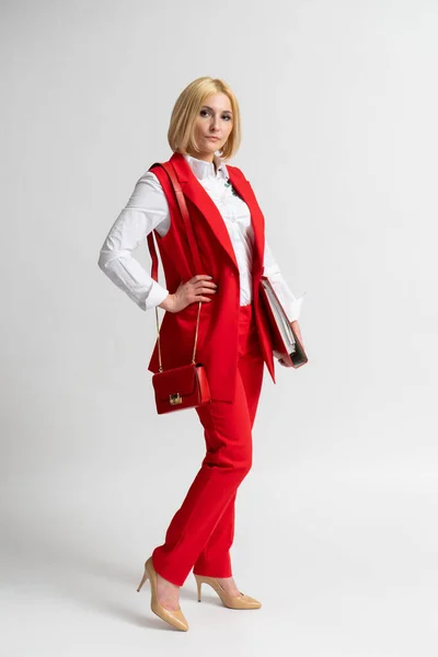 Young blonde business woman in a red casual trouser suit with a large office folder and a women\'s handbag on her shoulder posing on a white background