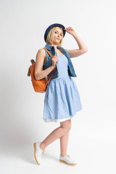 Happy young blonde woman in a blue casual summer dress, a denim vest with a backpack straightens her hat and poses in the studio on a white background.