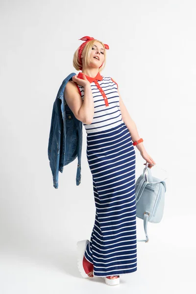 stock image Young blonde woman in a long casual striped dress with a red bandana on her head, a backpack, sunglasses and a denim jacket in her hand posing on a white background