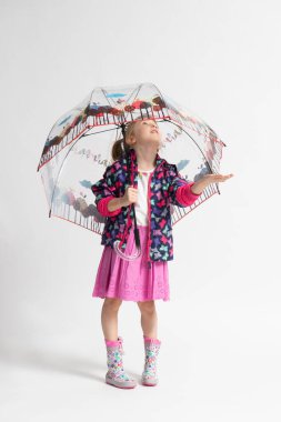 Pembe etekli, rahat ceketli ve lastik çizmeli küçük bir kız beyaz arka planda saydam renkli bir şemsiyenin altında yağmurdan saklanıyormuş gibi yapıyor.