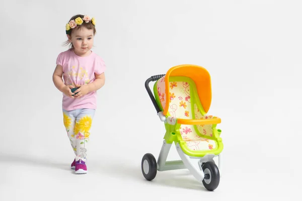 Beyaz arka planda oyuncak bebek arabasıyla oynayan küçük bir kız. Çocuk oyuncağı konsepti, çocukluk.