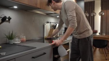 Bir adam evdeki mutfağı temizliyor, indüksiyon ocağını ve tezgahı temizleme çözeltisi ve temizleme beziyle dikkatlice siliyor..