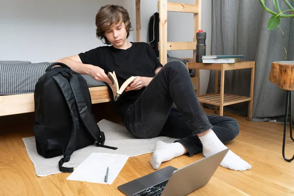 Genç bir öğrenci ya da öğrenci odasındaki ranzanın yanında dizüstü bilgisayar ve açık bir kitapla oturmuş ödev yapıyor, bir kağıt parçasına kalemle notlar yazıyor..