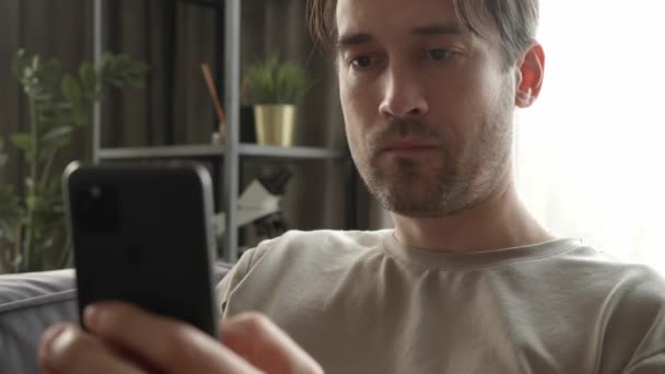 男性はスマートフォンの画面に無関心に見え 自宅でコンテンツをスクロールします ソーシャルネットワーク上の娯楽コンテンツの絶え間ない視聴からのドーパミン中毒の兆候 — ストック動画