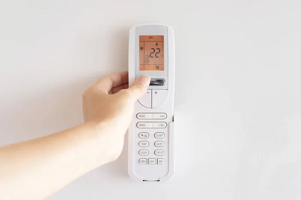A woman\'s hand turns on air conditioner remote control mounted on white wall. LCD display with orange backlight shows room temperature of 22 degrees Celsius and automatic cooling mode.