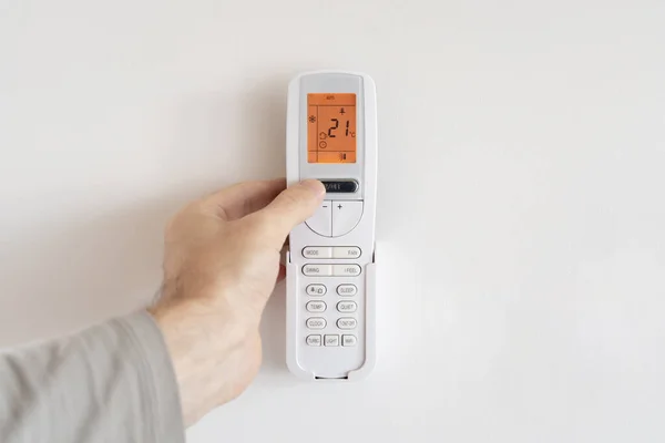 Male hand turns on air conditioner remote control mounted on white wall. LCD display with orange backlight shows room temperature of 22 degrees Celsius and automatic cooling mode.