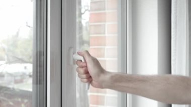 Adam plastik pencere çerçevesinin sapını eliyle çeviriyor ve odayı havalandırmak, temiz hava almak ve içerideki sıcaklığı değiştirmek için PVC penceresini hafifçe açıyor..