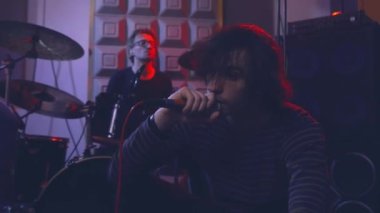 Bir şarkıcı duygusal olarak mikrofona şarkı söylüyor, bir müzik stüdyosunun zemininde hoparlörlerin yanında oturuyor ve davulcunun çaldığı bir davul takımı var..