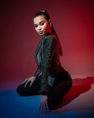 Genç ve güzel bir kadın kameraya poz veriyor. Stüdyoda dizlerinin üzerinde oturuyor. Kırmızı arka planda.