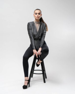 Yüksek topuklu ayakkabılar ve kameraya bakan göz kamaştırıcı bir kıyafeti olan güzel bir kız stüdyoda beyaz bir arka planda bir bar taburesinde zarifçe poz veriyor..