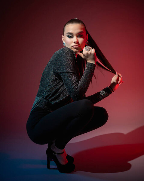 A young beautiful woman in high-heeled shoes poses looking at the camera, squatting on the floor in the studio on a red background