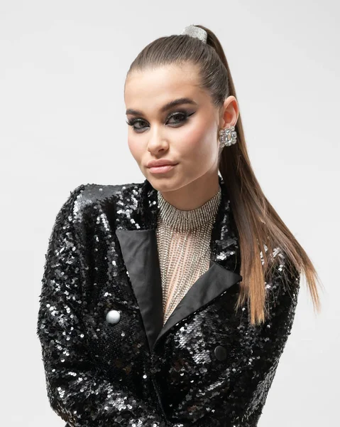 A model girl in a shiny jacket, with makeup, hairstyle and spectacular jewelry poses looking at the camera in the studio on a white background