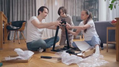 Ebeveynler ve çocukları yerde otururken yeni mobilyaların nasıl monte edileceği bilmecesini çözmek için birlikte çalışıyorlar. Bazı parçaların talimatlarına göre mobilyaların kendiliğinden montajı.