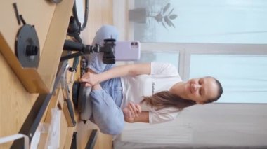 Kız video bağlantısıyla mobilyaların kendiliğinden montajı için bir takım parçaları ve aletleri gösteriyor, odanın içinde önünde uzanmış, bir tripoda monte edilmiş telefon kamerasını kullanıyor. Dikey çekim.