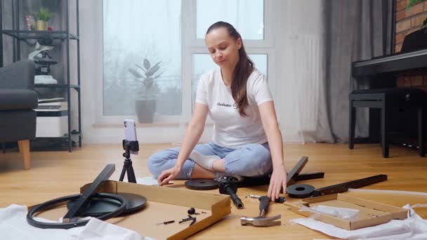 ブロガーの女の子は 彼女が自宅で家具を組み立てるときに使用するツールを電話カメラを通じて彼女の聴衆を示しています 女性は彼女のチャンネルのためにカメラでDiyビデオコンテンツを記録します — ストック動画