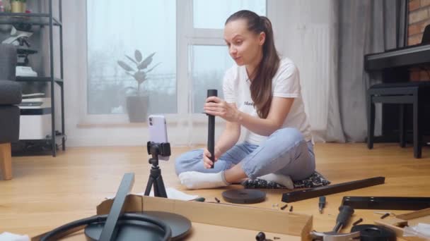女孩通过视频链接展示了一套家具自组装的零件和工具 这些零件和工具放在她面前的房间地板上 用她安装在三脚架上的电话摄像头 — 图库视频影像