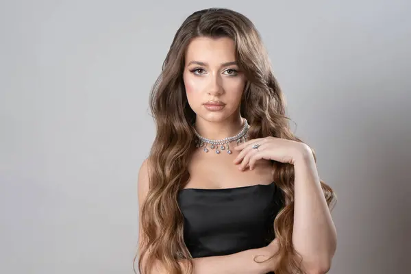 A beautiful long-haired girl in a black evening dress with a diamond ring on her finger and a necklace of pearls and precious stones around her neck, looking at the camera, poses in the studio.