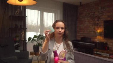 Islak saçlı bir kadın vlogger kameraya bigudileri gösteriyor ve elinde tarak, saç spreyi, saç köpüğü ve saç şekillendirme ürünleri şişeleri tutuyor. Saç stili kursu.