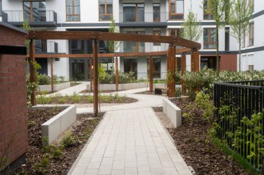 Ahşap pergola, peyzaj bahçeleri ve düzgün asfalt bir yolu olan modern bir konut avlusu. Arka plandaki çağdaş apartman binaları, sakin açık alanı tamamlıyor..
