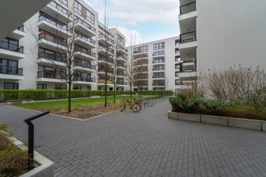 Temiz mimari çizgilere sahip modern bir apartman kompleksi, içinde genç ağaçlar ve bisiklet parkı olan manzaralı bir avlu. Şehir yaşamı ve yeşilliğin karışımı bir ortam sunuyor..