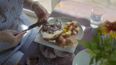 Bir kadın kahvaltıdan sonra tatlı olarak kruvasana çikolata ezmesi sürer. Tabağında meyve salatası ve yakınlardaki masada bir bardak su var..
