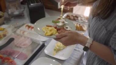 Bir kadın, otel büfesinde kendisine mozarella, domates aperatifi, omlet ve sosis ile kahvaltı tabağı servis ediyor. Otel restoranında self servis kahvaltı büfesi..