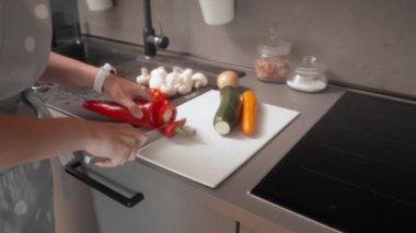 Bir kadın modern bir mutfakta beyaz bir kesme tahtasıyla kırmızı bir dolma biberi dilimliyor. Tahtanın etrafında bir kabak, havuç, mantar, kiraz domatesleri ve bir soğan da dahil olmak üzere taze sebzeler bulunuyor..
